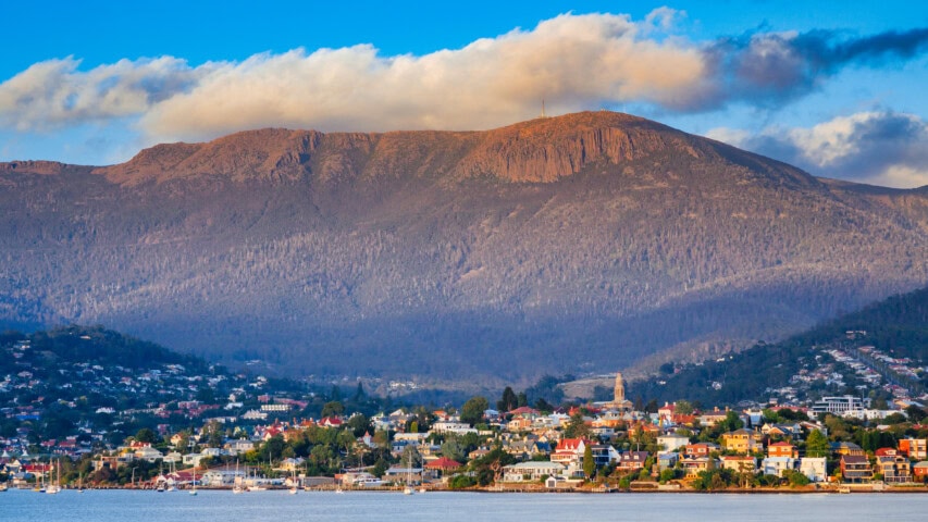 A coastal town in Tasmania with colorful houses sits at the base of a large, rocky mountain under a blue sky with clouds, making it a perfect travel destination for those seeking picturesque views and serene vibes.