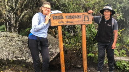 Two hikers stand next to a wooden sign for Montana Waynapicchu, which states the altitude and other information. Surrounded by greenery at this high-elevation location, they appear to be on an unforgettable journey through the Inca Trail towards the breathtaking vistas of Machu Picchu.