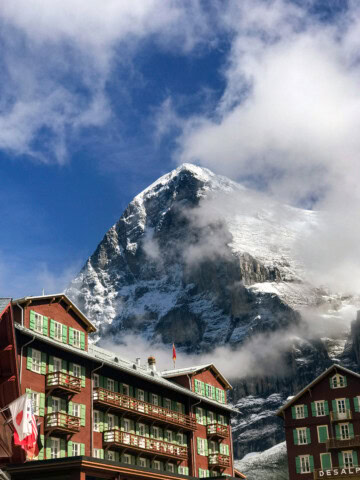 A snow-capped mountain in the Swiss Alps, partially obscured by clouds, is seen behind brown buildings with green shutters and balconies. A red flag with a white cross, symbolizing Switzerland, is flying on one of the buildings.