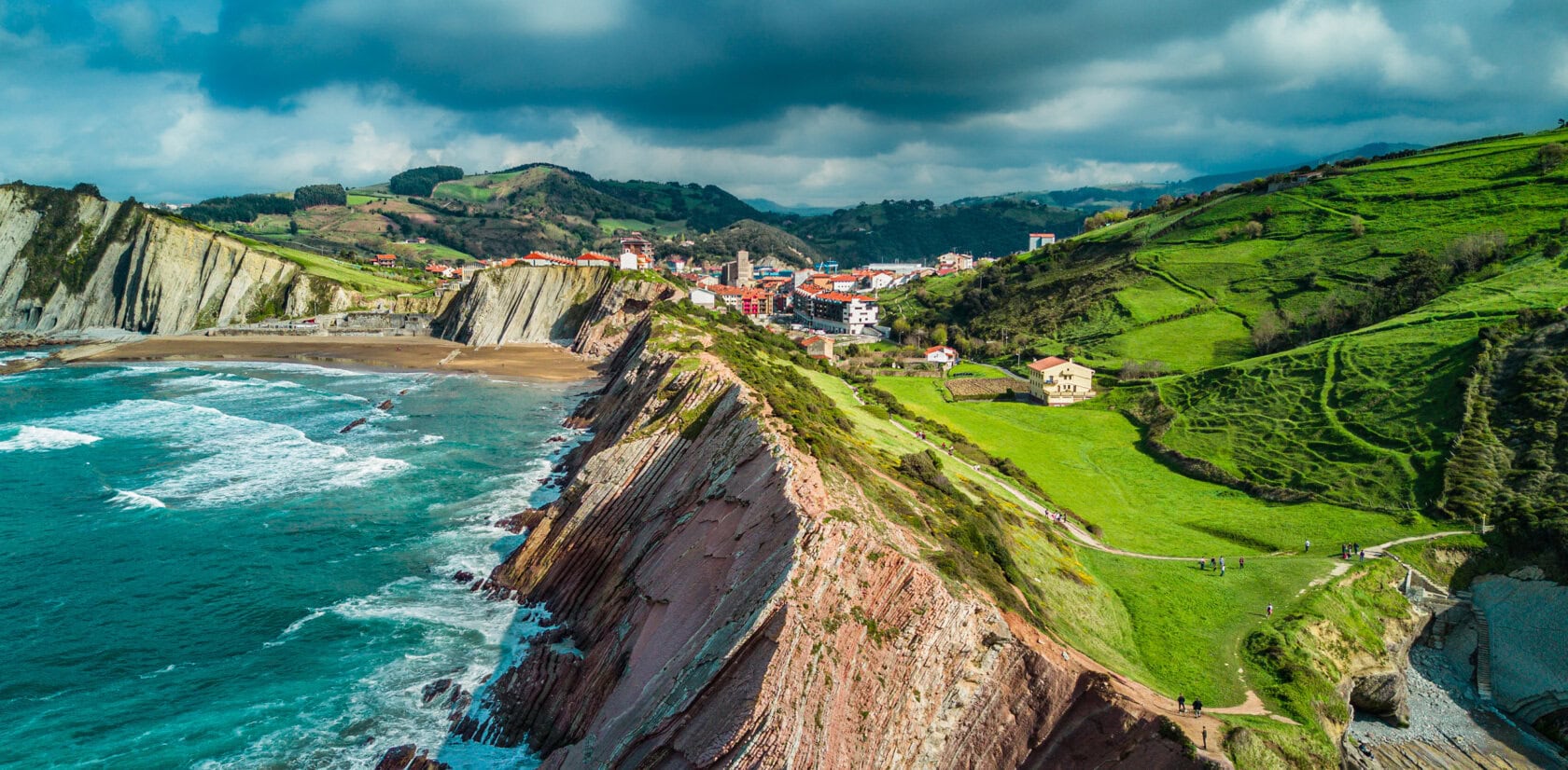 Aerial view of a coastal village with dramatic cliffs, green fields, and waves crashing against the rocky shoreline under a partly cloudy sky—an inspiring scene among the Top Ten Destinations for Travel 2026.
