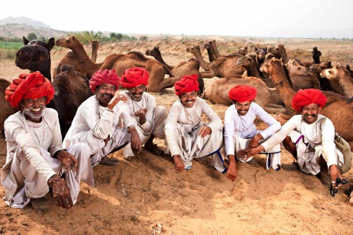 A group of five men wearing white garments and red turbans sit on the ground in front of a herd of camels, capturing the essence of a traveler's adventure in a sandy, arid landscape.