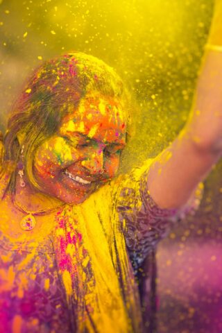 A traveler, smiling with closed eyes, is covered in colorful powder during a celebration, with yellow powder prominently in the air around them.