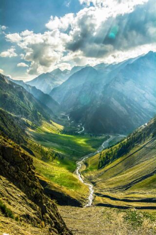 A lush green valley with a winding river is surrounded by tall mountains under a partially cloudy sky with sun rays breaking through, beckoning any traveler seeking adventure.