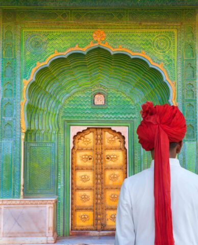 A person wearing a white garment and red turban stands facing an ornate, green arched entrance with an intricately designed golden door, ready to embark on their next adventure.