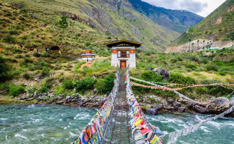 A suspension bridge adorned with colorful prayer flags spans across a river, leading to a white building amidst Bhutan's lush, mountainous landscape – a serene destination perfect for travel and tourism.