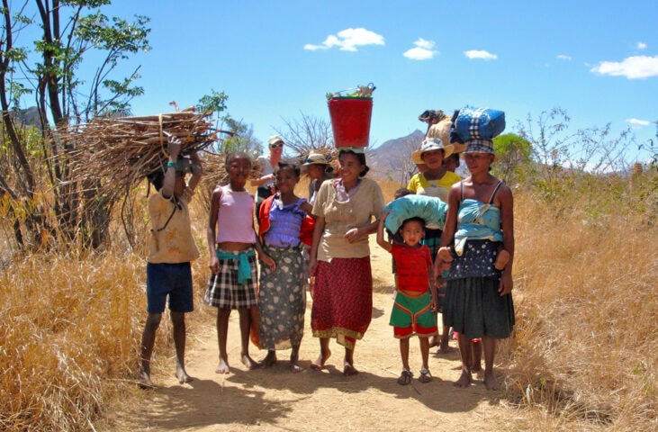 A group of people walking on a dirt path through The Eighth Continent's dry, grassy terrain. Some carry items on their heads, and one person shoulders a bundle of sticks. Trees and hills punctuate the background.