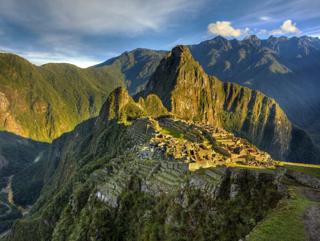 A panoramic view of Machu Picchu, Peru, showcasing ancient Incan ruins atop a mountain ridge surrounded by lush green hills and distant peaks under a blue sky with scattered clouds—an awe-inspiring sight that draws visitors year-round, regardless of the months.