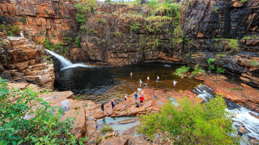 People are gathered on rocky terrain beside a waterfall and natural pool surrounded by cliffs and lush greenery, experiencing the pristine beauty that Australia has to offer.