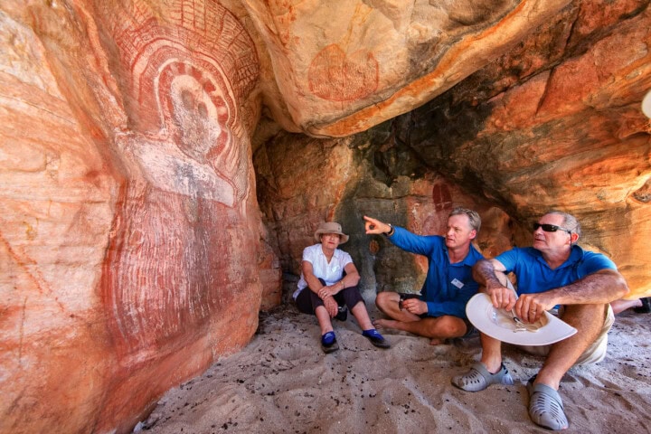 Three people observe and discuss ancient rock art in a cave in Australia. One person points towards the painted figure on the textured cave wall. The cave features sandy ground and rugged rock surfaces.
