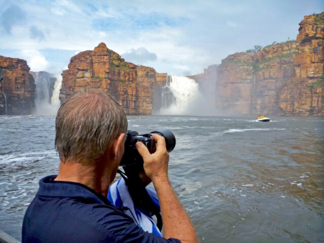 A person photographs a large waterfall cascading between rocky cliffs in Australia, with a speedboat visible in the water nearby.