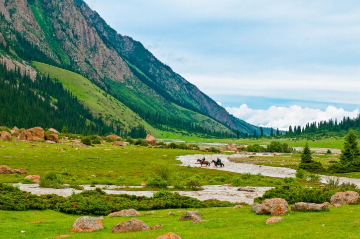 Three people on horseback ride through a green valley with a rocky riverbed, surrounded by mountains and trees under a partly cloudy sky in Kyrgyzstan.
