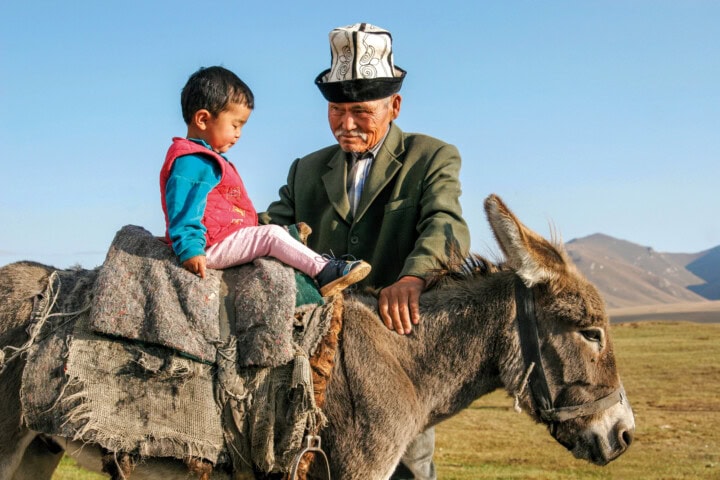 An older man wearing a hat helps a young child sit on a donkey, with the rugged mountains of Kyrgyzstan in the background.