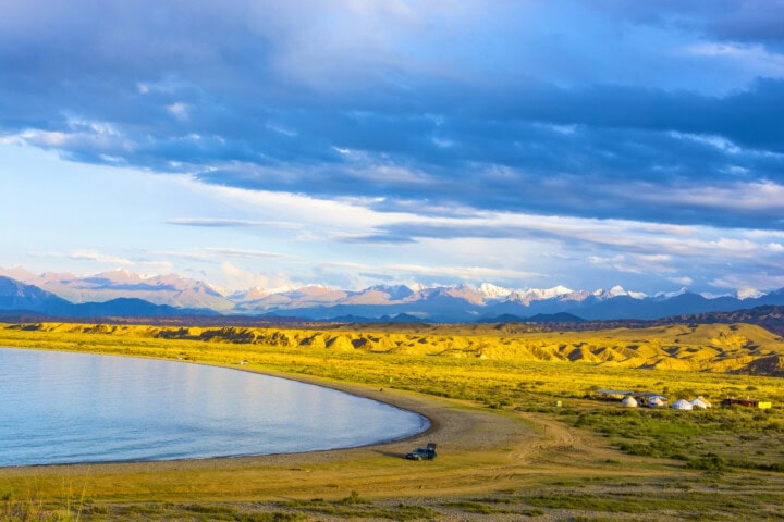 A scenic view of a calm lake bordered by a sandy shore in Kyrgyzstan, with a vast, yellow grassland transitioning into distant snow-capped mountains under a partly cloudy sky. A vehicle and tents are visible near the shore.
