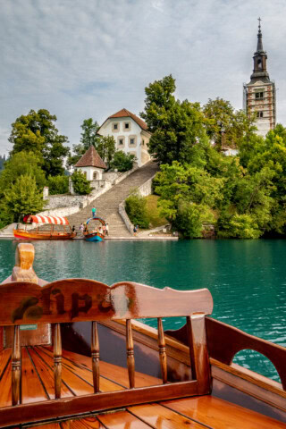 A traditional wooden boat on turquoise waters approaches a small island with a white church and a bell tower surrounded by greenery, offering an idyllic travel experience in Slovenia.
