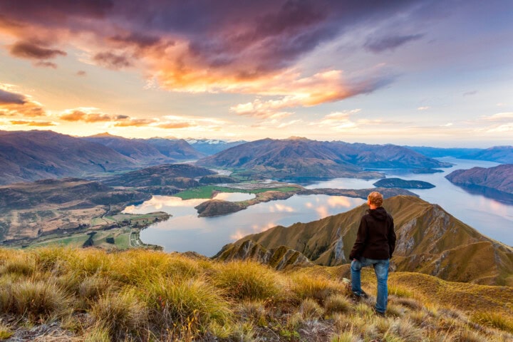 A person stands on a grassy hillside in New Zealand, overlooking a scenic view of a lake, mountains, and a dramatic sky at sunset.