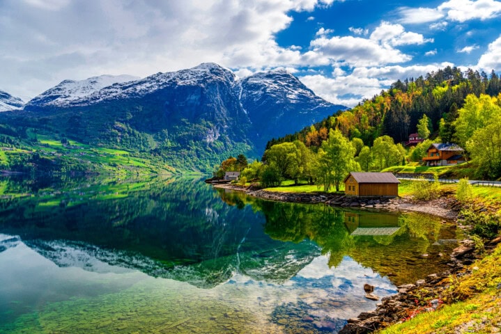 A tranquil lake in Norway reflects snow-capped mountains and a hillside with green trees and cabins under a partly cloudy sky.