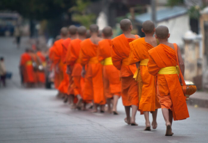 A group of monks with shaved heads and wearing bright orange robes walk in a single file along a street in Laos.