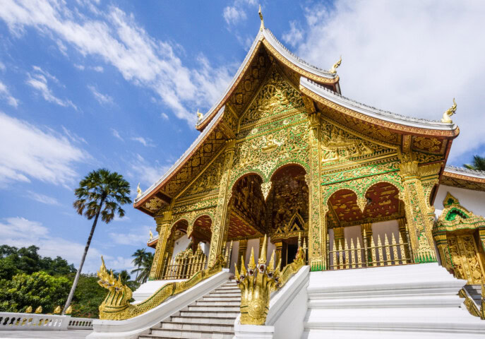Ornate temple with golden details and a multi-tiered roof, flanked by palm trees under a blue sky. White steps lead up to the entrance, which features intricate carvings and decorations.