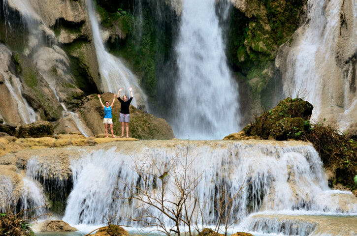 Two people stand with raised arms on a rocky ledge in front of a cascading waterfall amidst lush greenery, showcasing the breathtaking beauty of Laos.