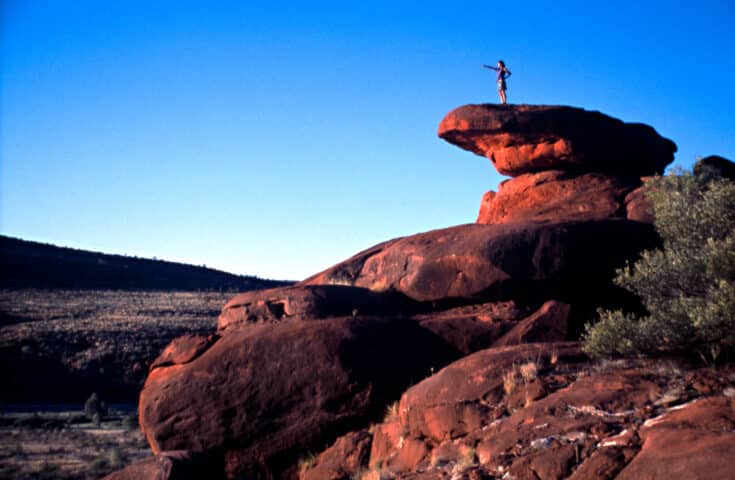 A person stands atop a large, red rock formation against a clear blue sky in Australia, seemingly pointing into the distance. The arid landscape below, dotted with some vegetation, enhances the allure of this travel destination for tourism enthusiasts.