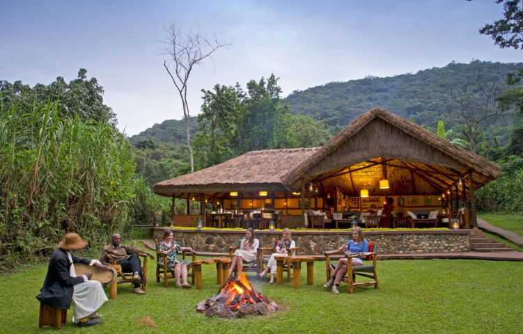 A group of people sit around a campfire on wooden chairs outside a rustic, thatched-roof lodge surrounded by lush greenery in a mountainous area, their adventure guided by the beauty of nature.