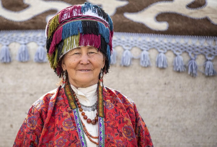 An elderly woman in traditional colorful attire and a fringed hat stands in front of a decorated wall in Kyrgyzstan, smiling.