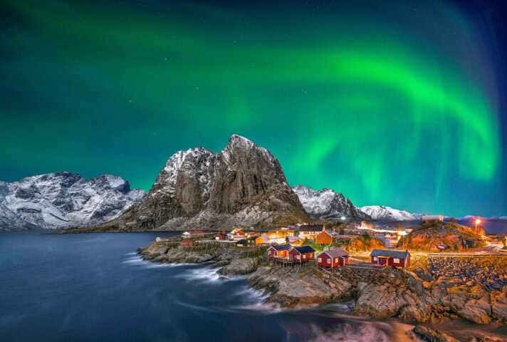 Coastal village in Norway with red houses, snow-covered mountains, and the aurora borealis illuminating the night sky.