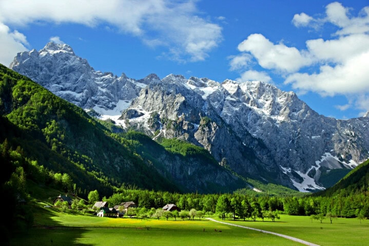 A serene landscape featuring a lush green valley, scattered houses, and a winding road, set against a backdrop of towering, snow-capped mountains under a blue sky with scattered clouds in picturesque Slovenia.