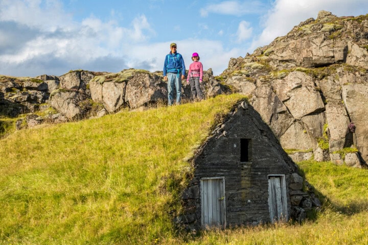 Two people stand on a grassy hillside in Iceland near a small rustic stone shed with two wooden doors, surrounded by rocky cliffs under a partially cloudy sky—an ideal scene for travel enthusiasts seeking authentic tourism experiences.