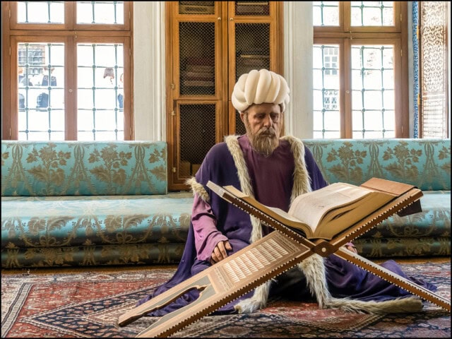 A person dressed in historical attire sits on a carpeted floor, reading an open book placed on a large wooden stand. The cozy room, reminiscent of Turkey’s rich history, features a couch and windows in the background.