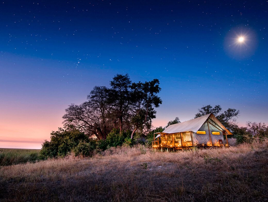 A lit canvas tent stands on a grassy hill at dusk, surrounded by trees, with a starry sky and bright moon overhead—the perfect end to a memorable hiking trip.