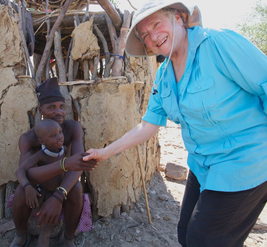 An elderly person in a blue shirt and hat shakes hands with a seated individual holding a child, set against the backdrop of a rustic, partially wooden structure that feels like something from an adventure in Namibia.