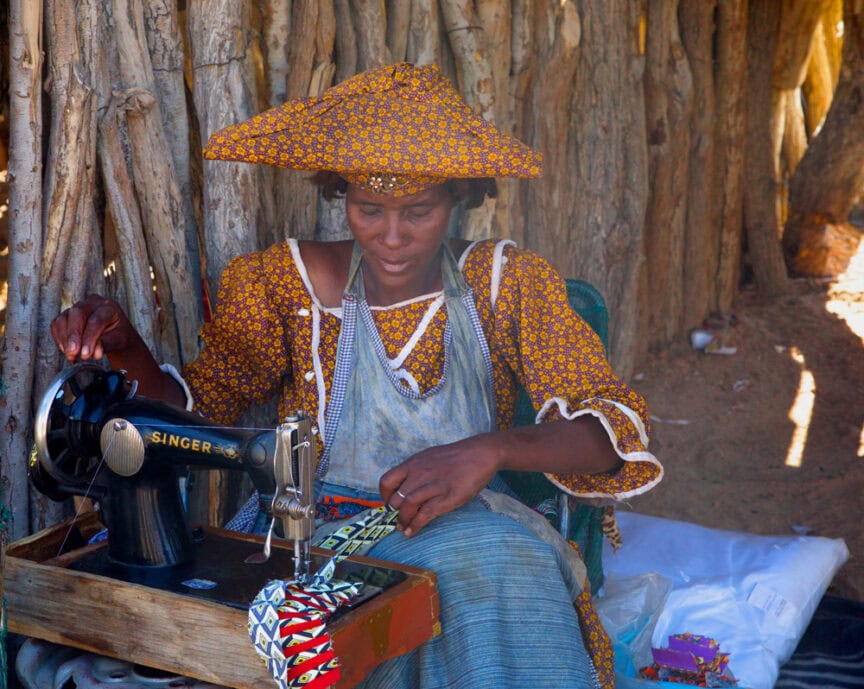 A person operates a vintage Singer sewing machine outdoors in Namibia, wearing a patterned hat and dress, adding a touch of Wonderland to their adventure.