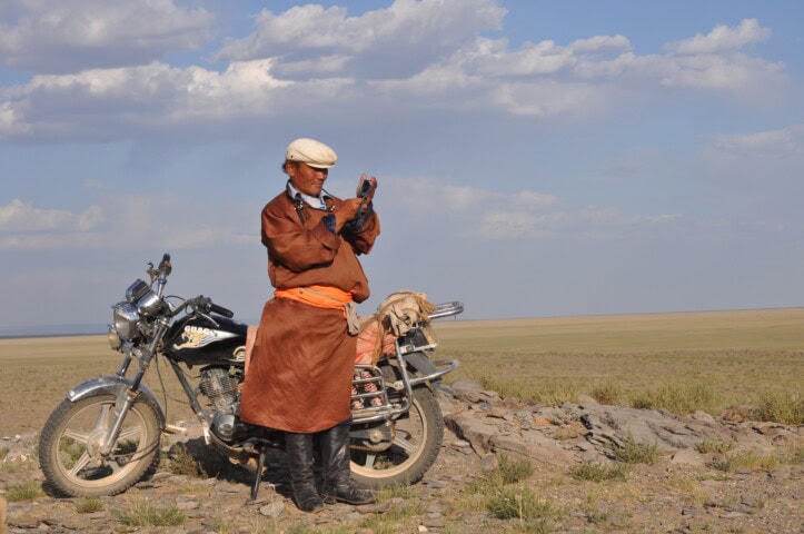 A person wearing traditional attire stands near a motorcycle in a vast, open landscape under a partly cloudy sky. They appear to be using a mobile device, embodying the spirit of travel and tourism in Mongolia.