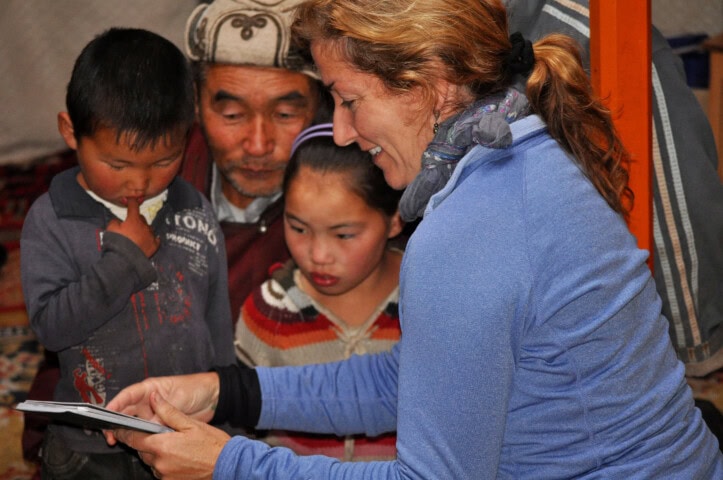 A woman shows a travel book to an elderly man and two children in a cozy indoor setting, sparking their curiosity about the distant landscapes of Mongolia.