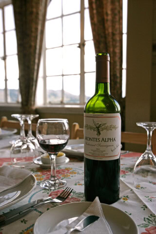 A bottle of Montes Alpha Cabernet Sauvignon sits on a table with a glass of red wine, plates, and silverware set for a meal, framed by curtains that bring to mind the elegance of Argentina.