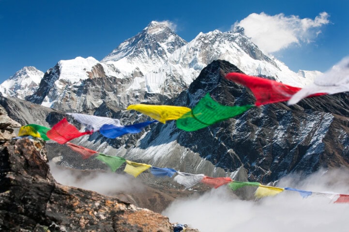 Prayer flags flutter in the foreground with a backdrop of Nepal's rugged mountains and snowy peaks under a clear blue sky.