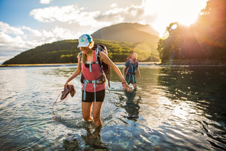 Two hikers wearing backpacks walk through shallow water at sunset with a mountain in the background. The front hiker holds her shoes in one hand, as they revel in the breathtaking beauty of New Zealand.