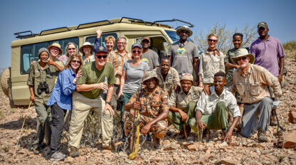 A group of 16 people, including tourists and wildlife conservationists, stand together in front of a safari vehicle on a rocky landscape in Namibia, smiling at the camera. Several hold walking sticks and binoculars, marking this special event with joy and camaraderie.