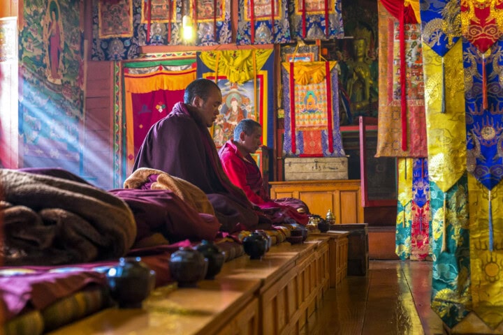 Two monks sit in a brightly decorated prayer room in Nepal, bathed in natural light pouring through the windows, surrounded by colorful banners and traditional decor.