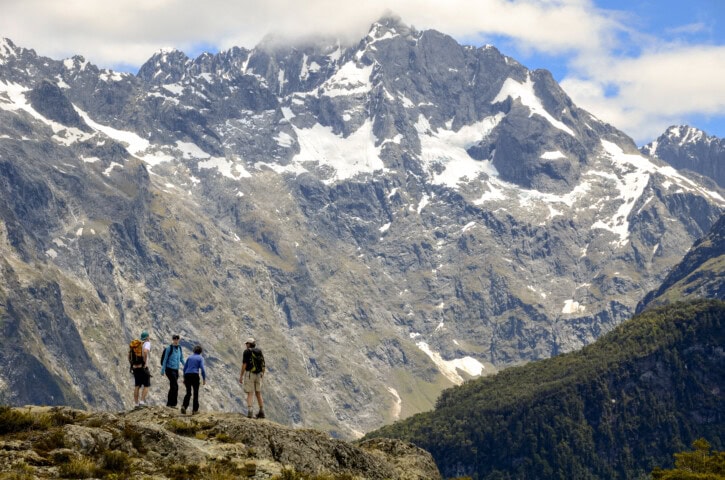 Four hikers stand on a rocky terrain in New Zealand, gazing at snow-capped mountain peaks under a partly cloudy sky.