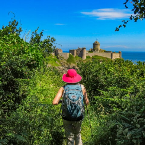 A person wearing a pink hat and carrying a backpack hikes on a path through greenery with a view of a castle in the background under the blue sky of France.