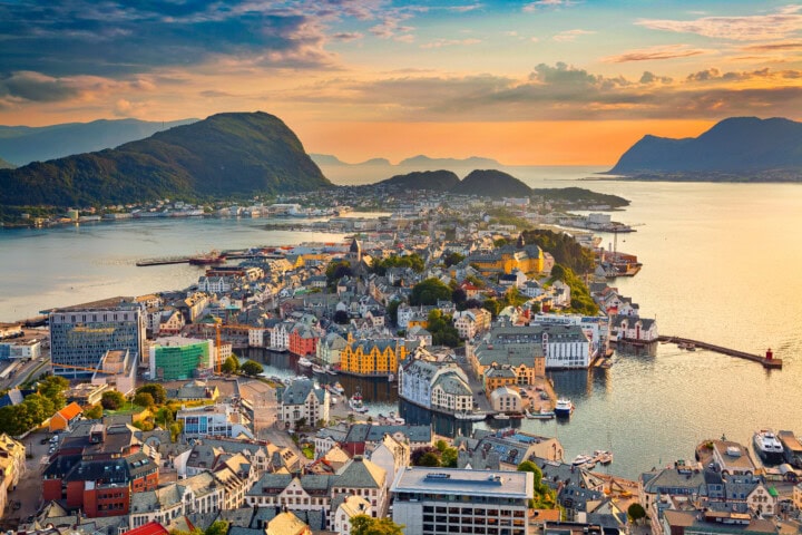 A scenic aerial view of Ålesund, Norway, at sunset. The town, a gem for tourism and travel enthusiasts, is surrounded by water with colorful buildings and majestic mountains in the background.