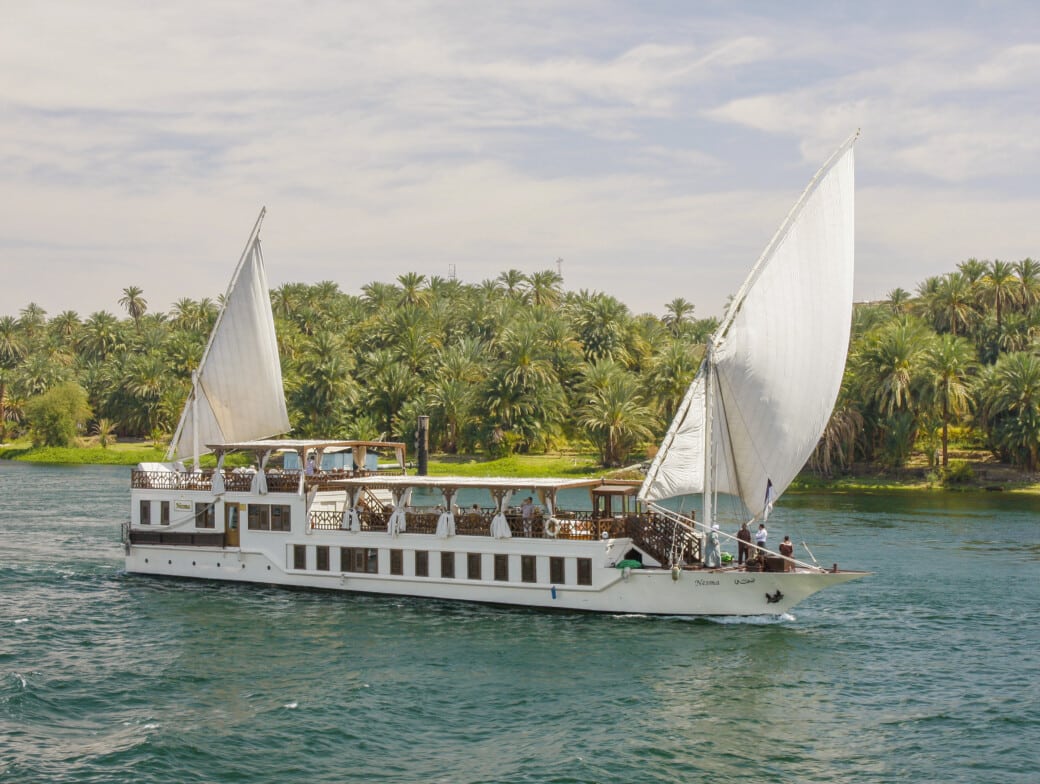 A white sailing boat with tall sails glides on a river, surrounded by palm trees and greenery on the riverbank under a partly cloudy sky, offering enchanting views all months.