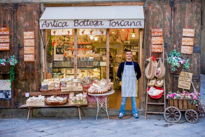 A shop owner stands in front of "Antica Bottega Toscana," a store with a rustic wooden facade, displaying a variety of traditional Italian goods and products outside, inviting travelers to experience the charm of Italy's rich heritage.