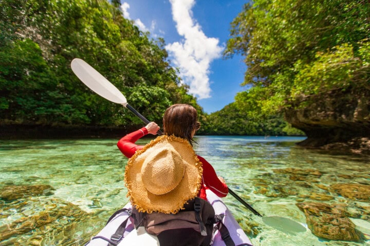 A person wearing a straw hat and red shirt kayaks through the clear, shallow waters of Palau, surrounded by lush greenery under a bright blue sky, embodying the perfect blend of adventure and travel.