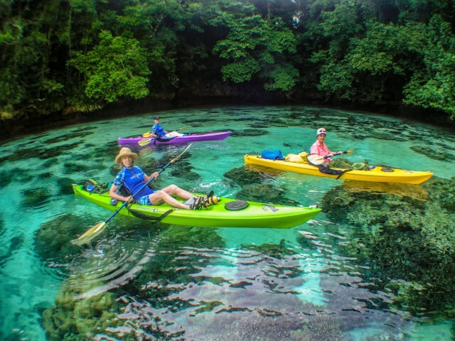 Three people kayaking in clear, turquoise water surrounded by lush greenery in the Pacific Islands; one person in a green kayak, another in a purple kayak, and the third in a yellow kayak.