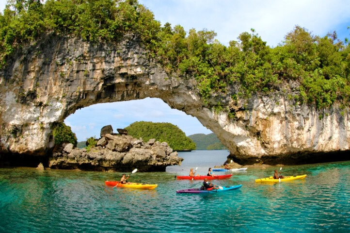 Kayakers paddle through clear turquoise waters under a natural rock arch, surrounded by lush greenery—a perfect snapshot of travel and tourism in Palau.
