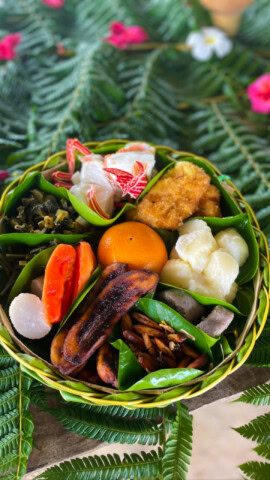 A beautifully arranged basket of various foods, featuring an orange, papaya slices, a fried item, white pieces, greens, and a banana on a bed of green leaves and fern fronds—a perfect snapshot of Palau's culinary delights for any travel enthusiast.