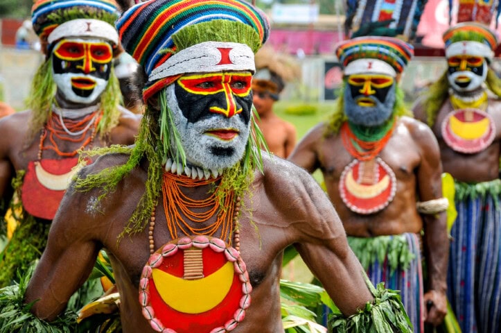 Men in traditional tribal attire with painted faces, colorful headdresses, and beaded necklaces participate in a cultural event outdoors in Papua New Guinea.